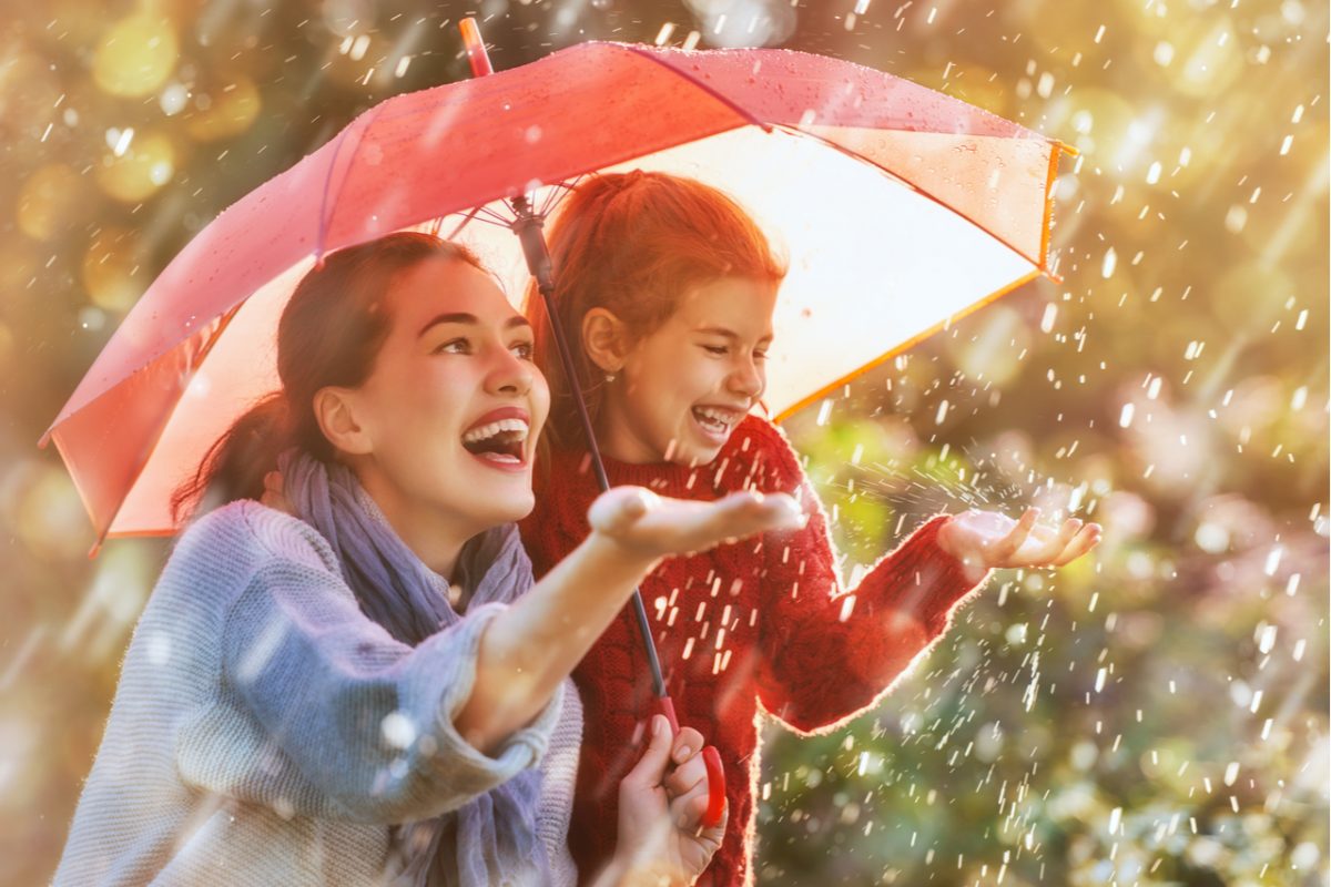 A mother and daughter laughing in the rain under a red umbrella.