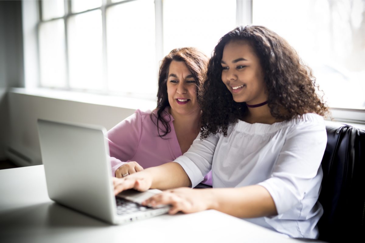 A happy mother and daughter sit at a desk working on a laptop together.