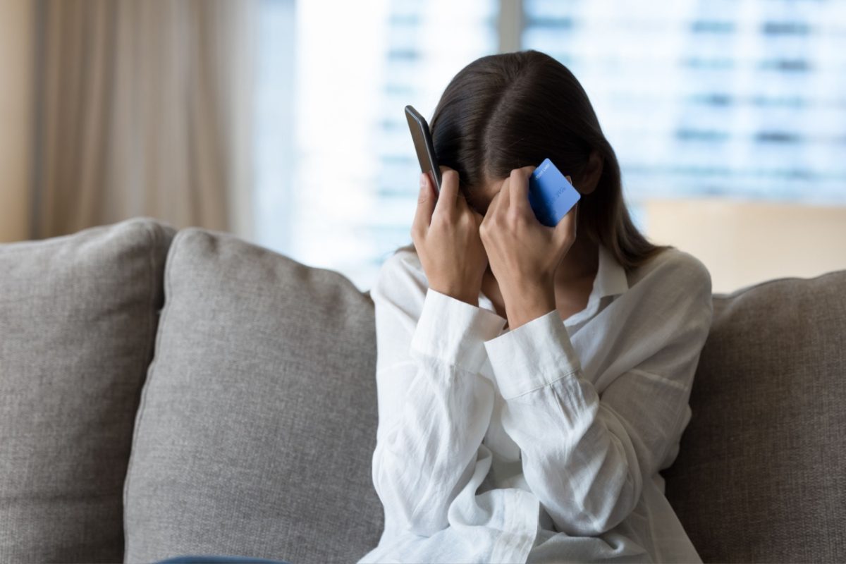 A woman holds her head with a phone in one hand and a bank card in the other.